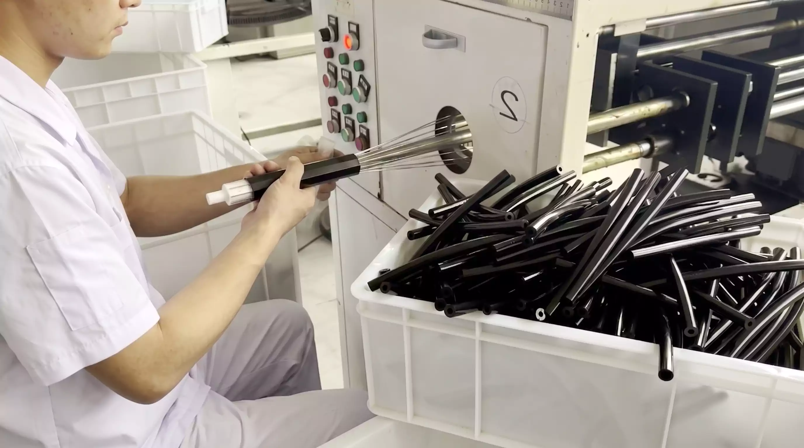 A manufacturing photo from a silicone cold shrink tube factory showing a worker in a white uniform using a machine to expand a black silicone sleeve onto a white spiral core. A bin of black tubes is on the left. A manufacturing photo from a silicone cold shrink tube factory showing a worker in a white uniform using a machine to expand a black silicone sleeve onto a white spiral core. A bin of black tubes is on the left.